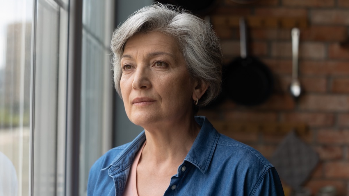 Elderly woman stands by window look away.