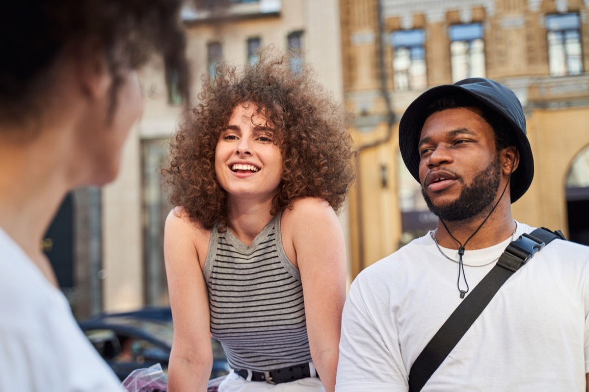 Smiling young man and woman are looking at curly female while walking in city centre