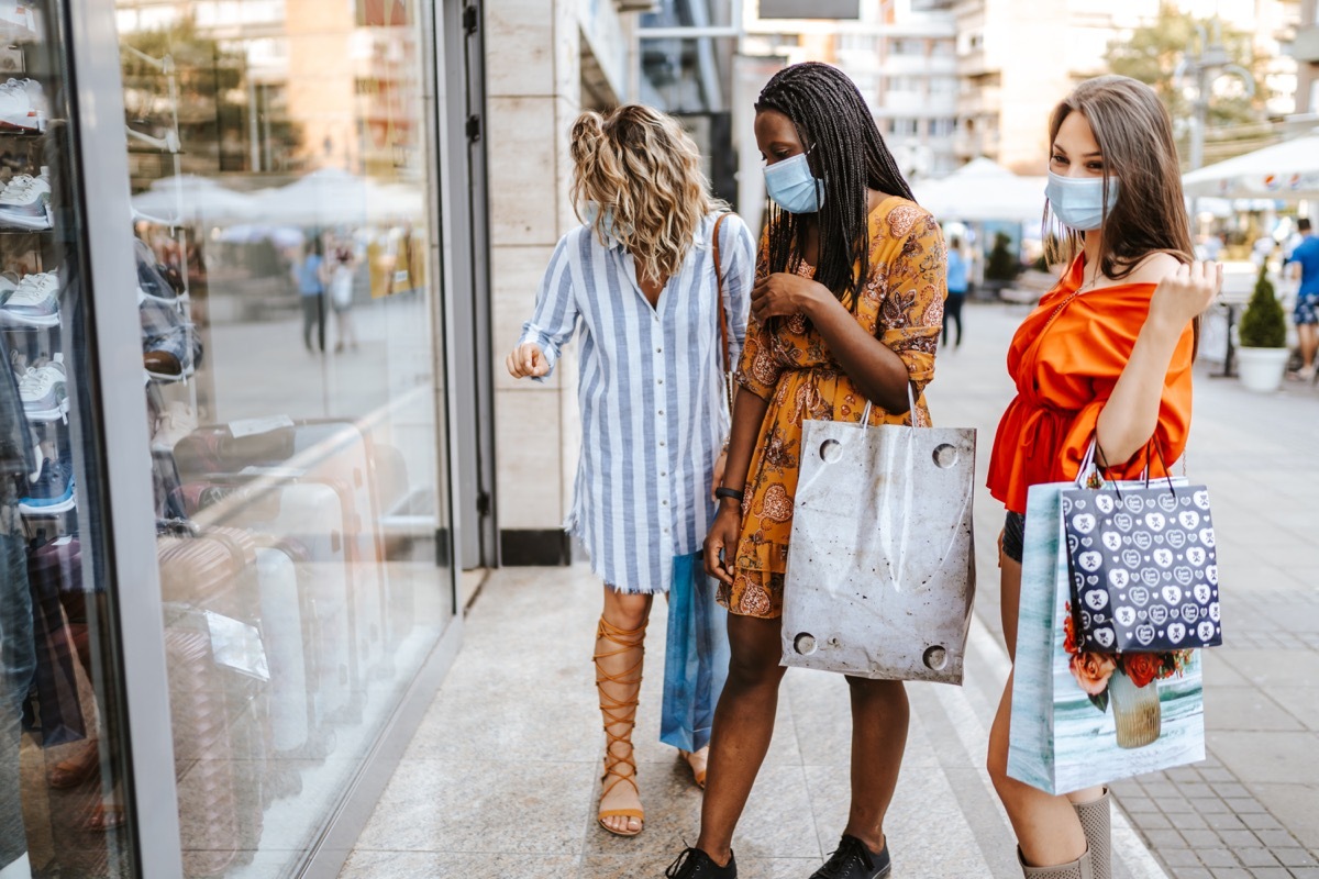 Three cheerful young friends wearing protective mask, walking down the street and shopping after reopening stores after COVID-19