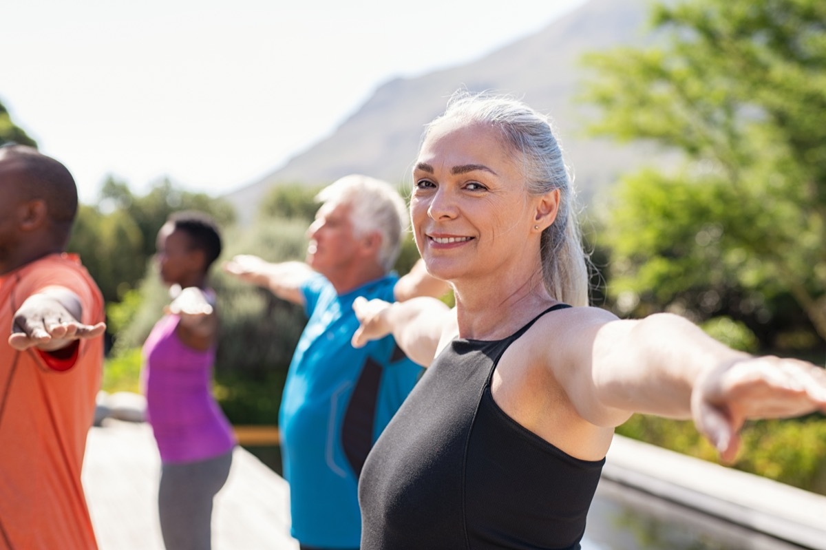 Portrait of happy senior woman practicing yoga outdoor with fitness class. Beautiful mature woman stretching her arms and looking at camera outdoor. Portrait of smiling serene lady with outstretched arms at park. (Portrait of happy senior woman pract