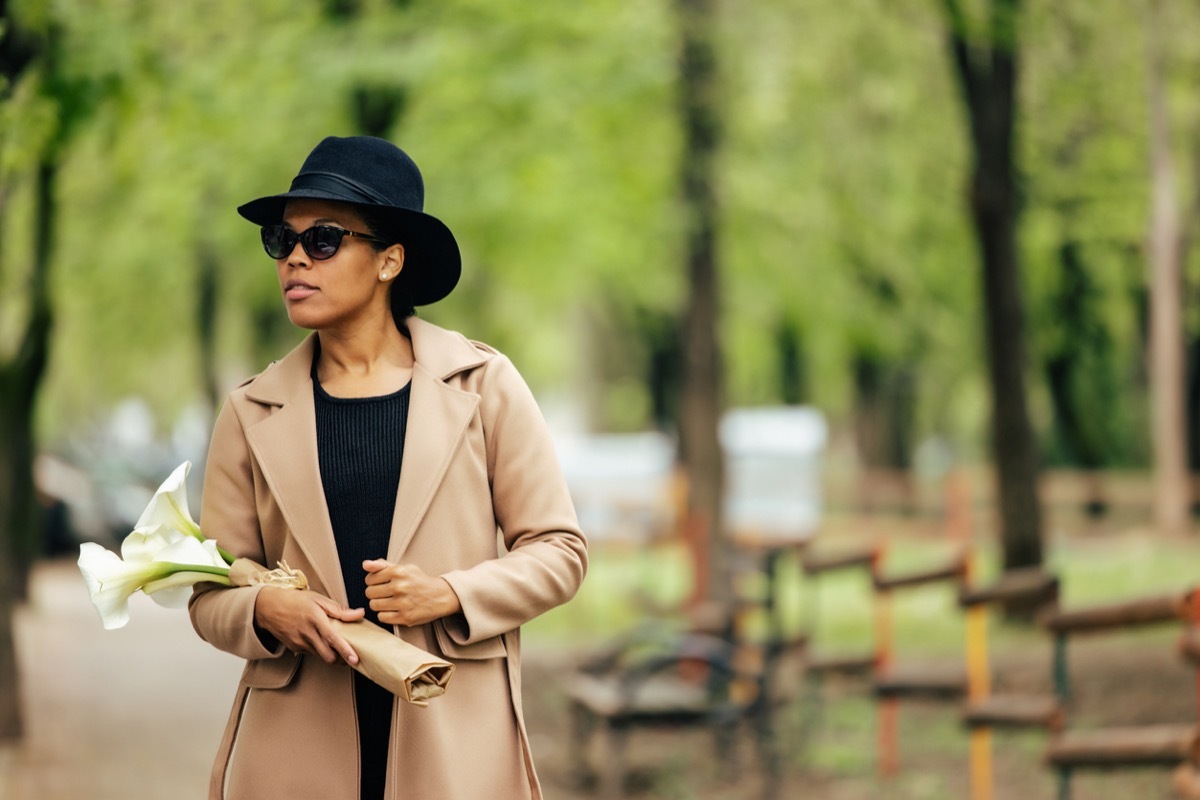 fashionable woman wearing camel coat and black fedora, carrying lilies while walking through the park