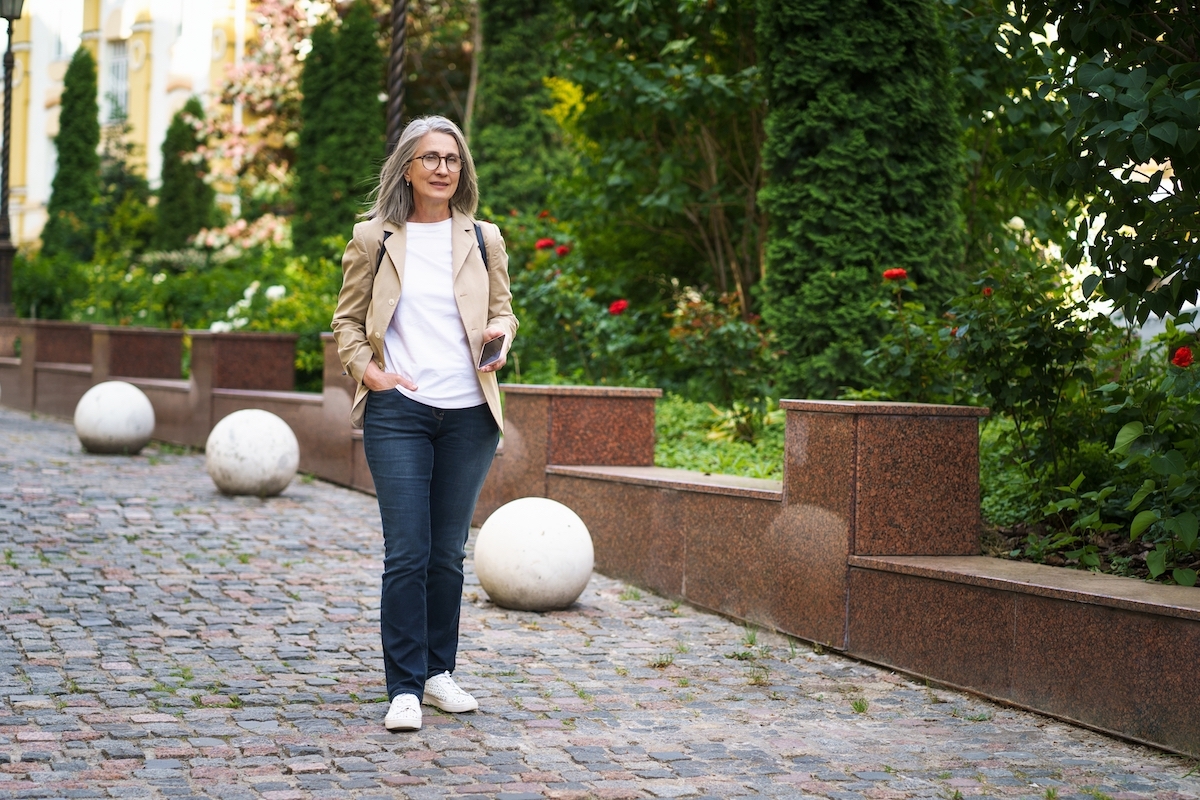 A mature woman wearing dark jeans, a white t-shirt, and a beige blazer walking down a pretty cobblestone street