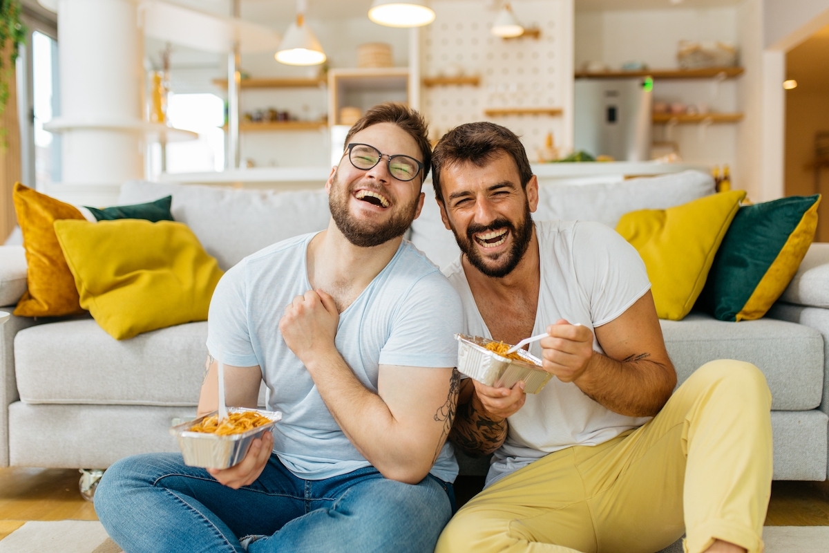 A male couple eating pasta out of takeout containers while sitting on their living room floor and laughing.