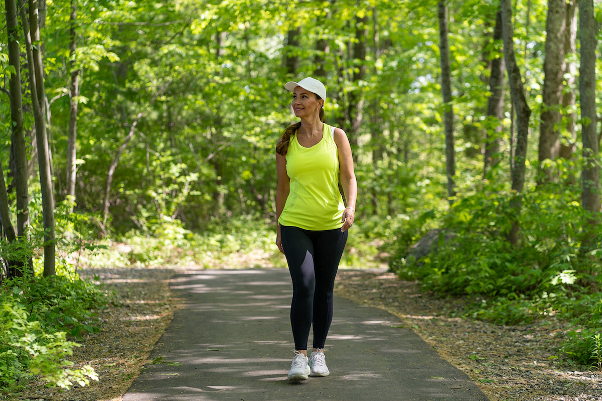 A middle-aged woman wearing black leggings and a bright yellow tank is taking a walk along a tree-lined path