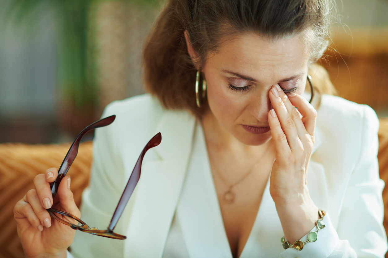 A middle-aged woman wearing a white blouse and jacket takes off her glasses to rub her eyes.