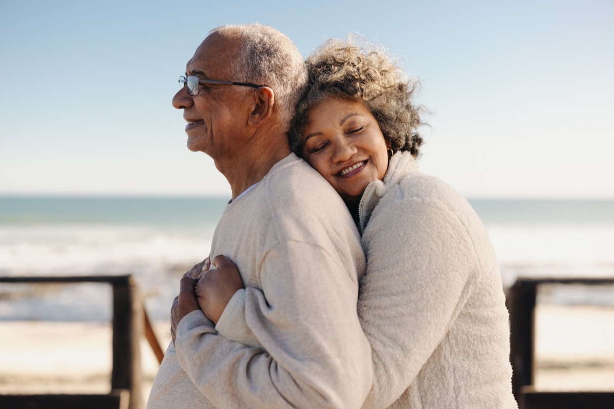 Affectionate senior woman smiling happily while embracing her husband by the ocean. Romantic elderly couple enjoying spending some quality time together after retirement.