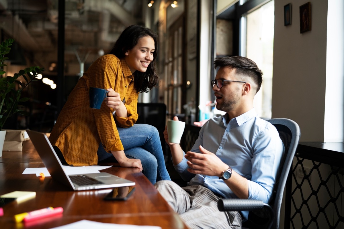 woman sitting on man's desk as they drink coffee