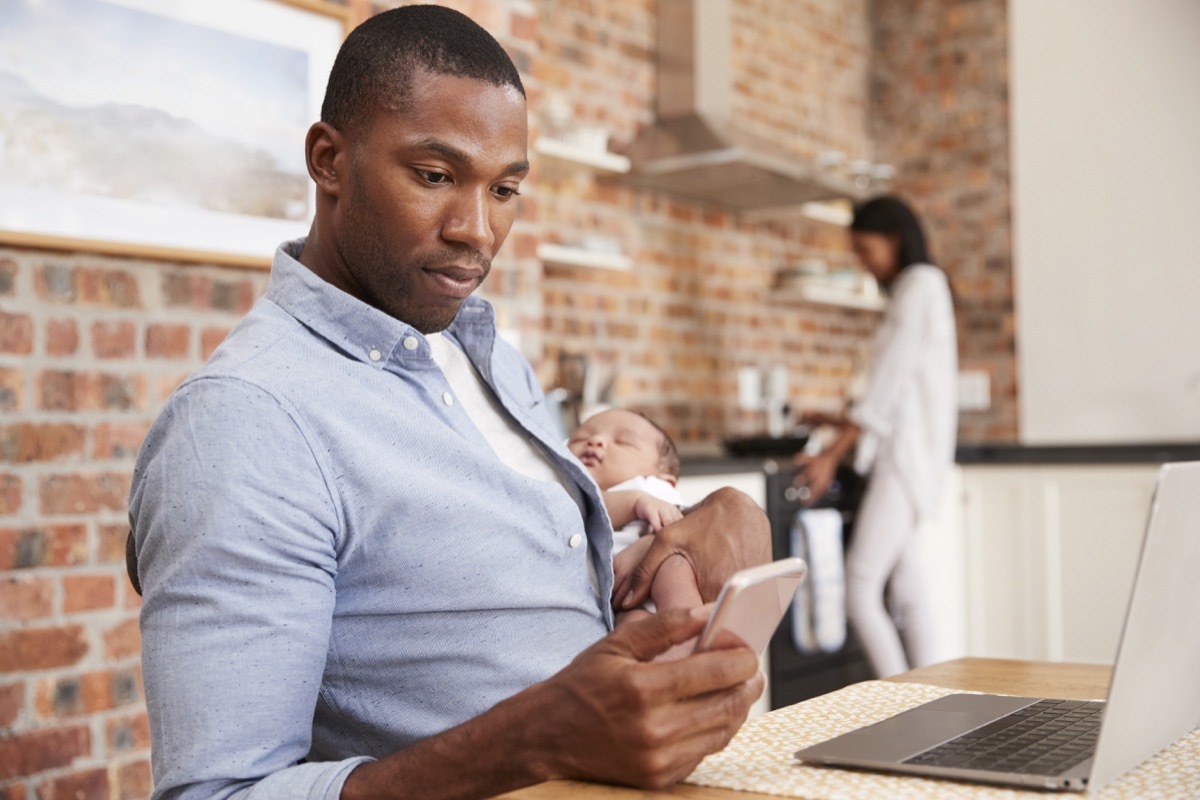 Father On Laptop Holds Newborn Son As Mother Makes Meal