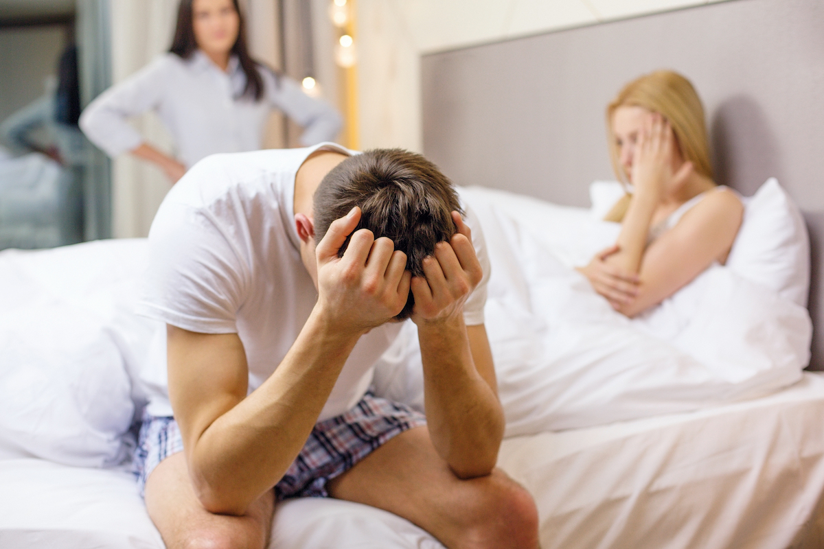 A man sitting on the bed with his head in his hands with a woman in the bed and another woman at the door, as he's been caught cheating.