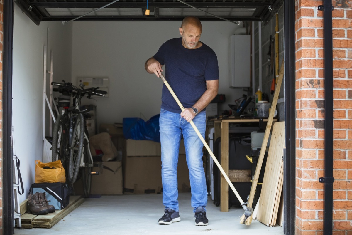 A man is sweeping the floor up his home garage with a large brush. The garage door is open.