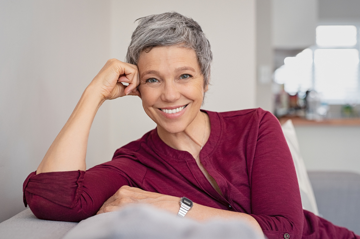 Smiling woman with short gray hair wearing a pink shirt and sitting on the couch.