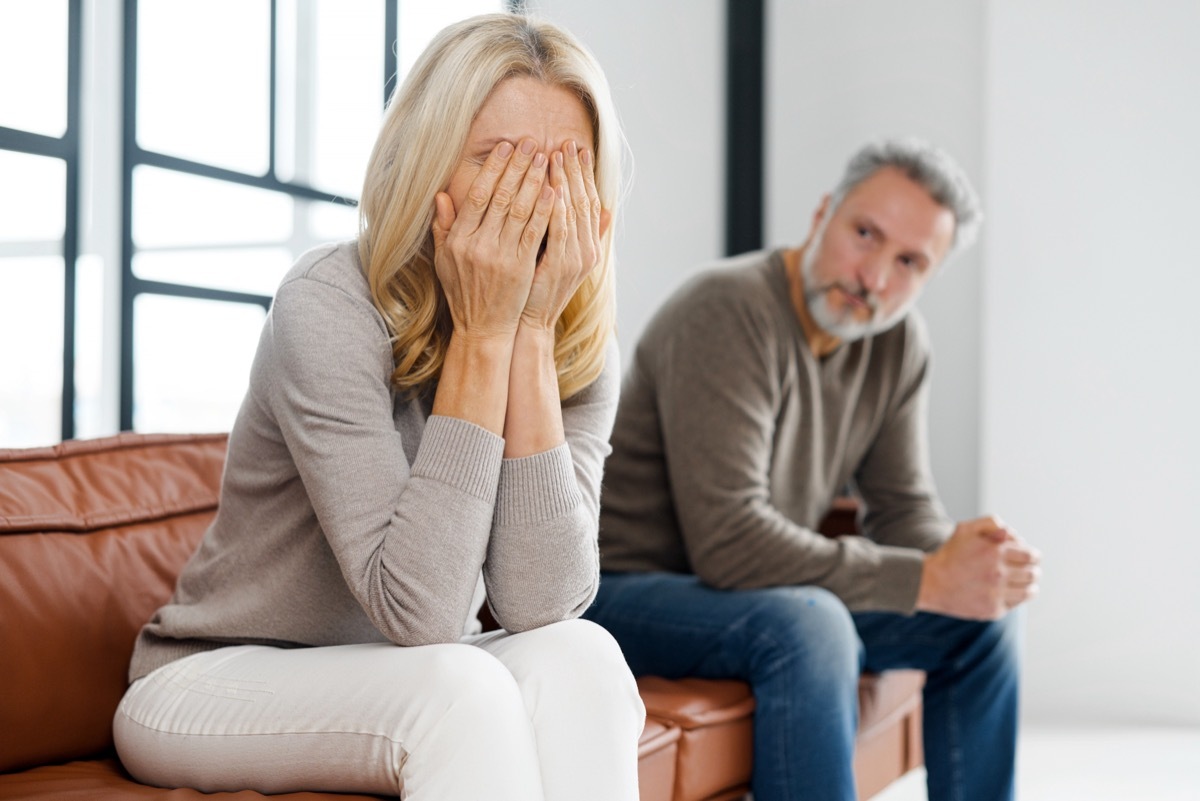 upset woman sitting on the couch with her head in her hands; a man looking on is in the background