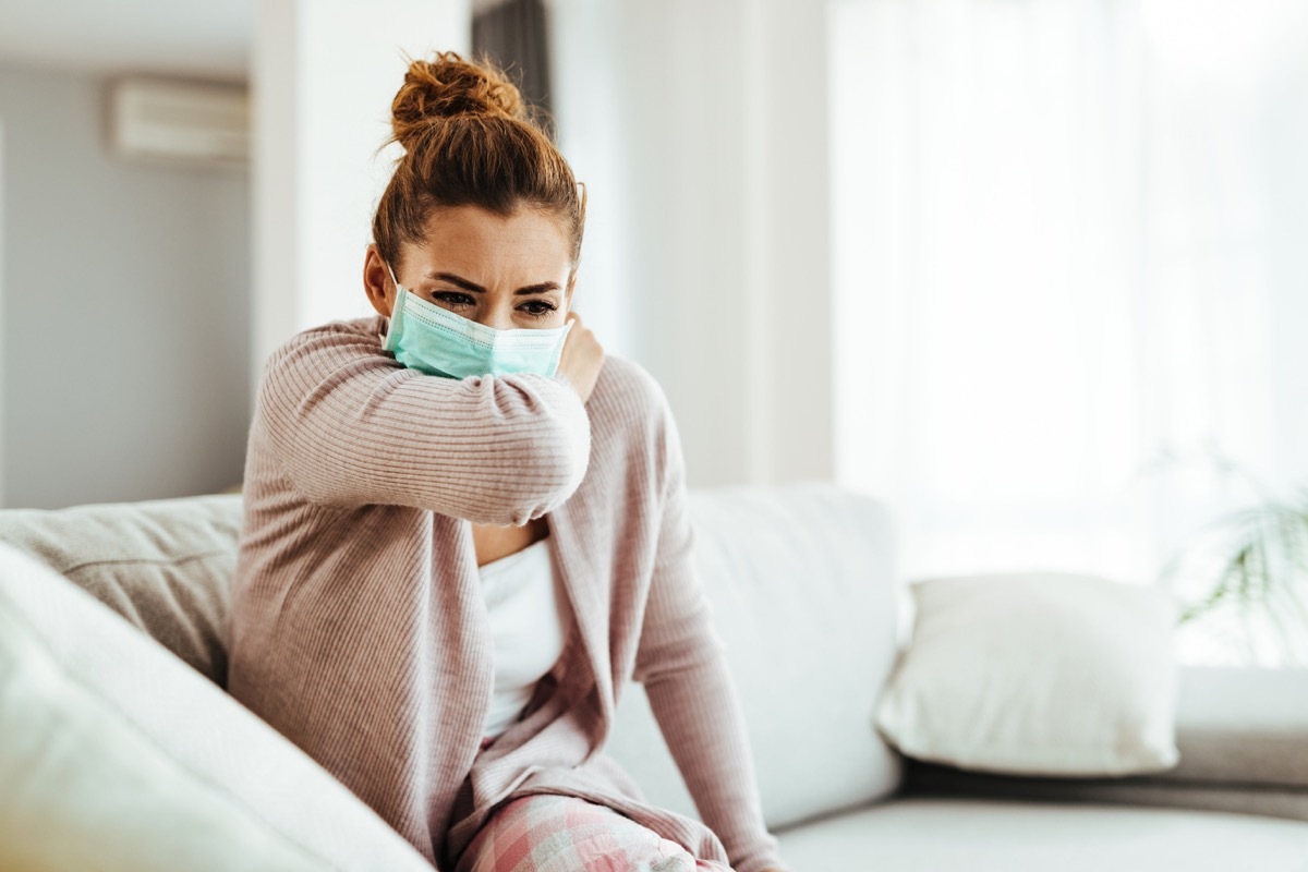 Woman wearing sneezing into her elbow while wearing protective face mask at home.