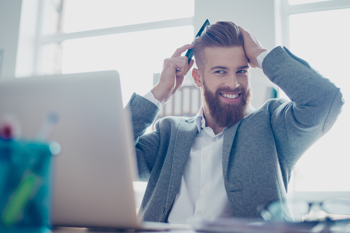 A young man smiling and combing his hair in front of his laptop at his desk in the office.