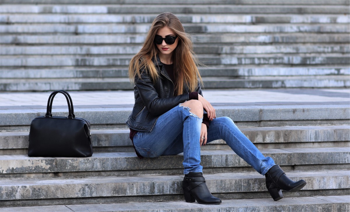 Casual style. Stylish woman in sunglasses sitting on the stone stairs. She dressed in leather jacket, skinny jeans and shoes on heels. Her sight directed downwards. Long hair, elegant, street fashion