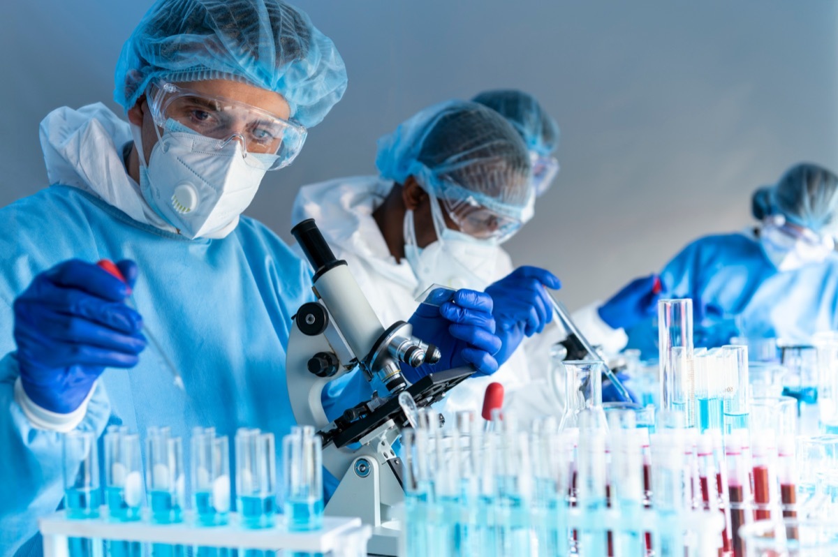 Virus experts are working using microscope in laboratory with protective suit, gloves glasses and mask