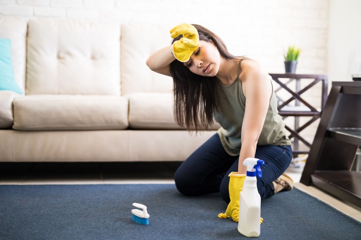 Young woman rubbing head wearing casuals and yellow gloves cleaning carpet with liquid soap