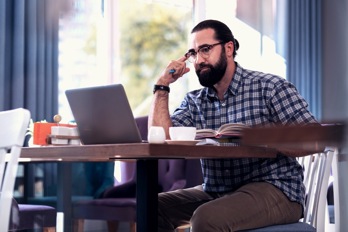 man wearing glasses sitting at table with book and laptop