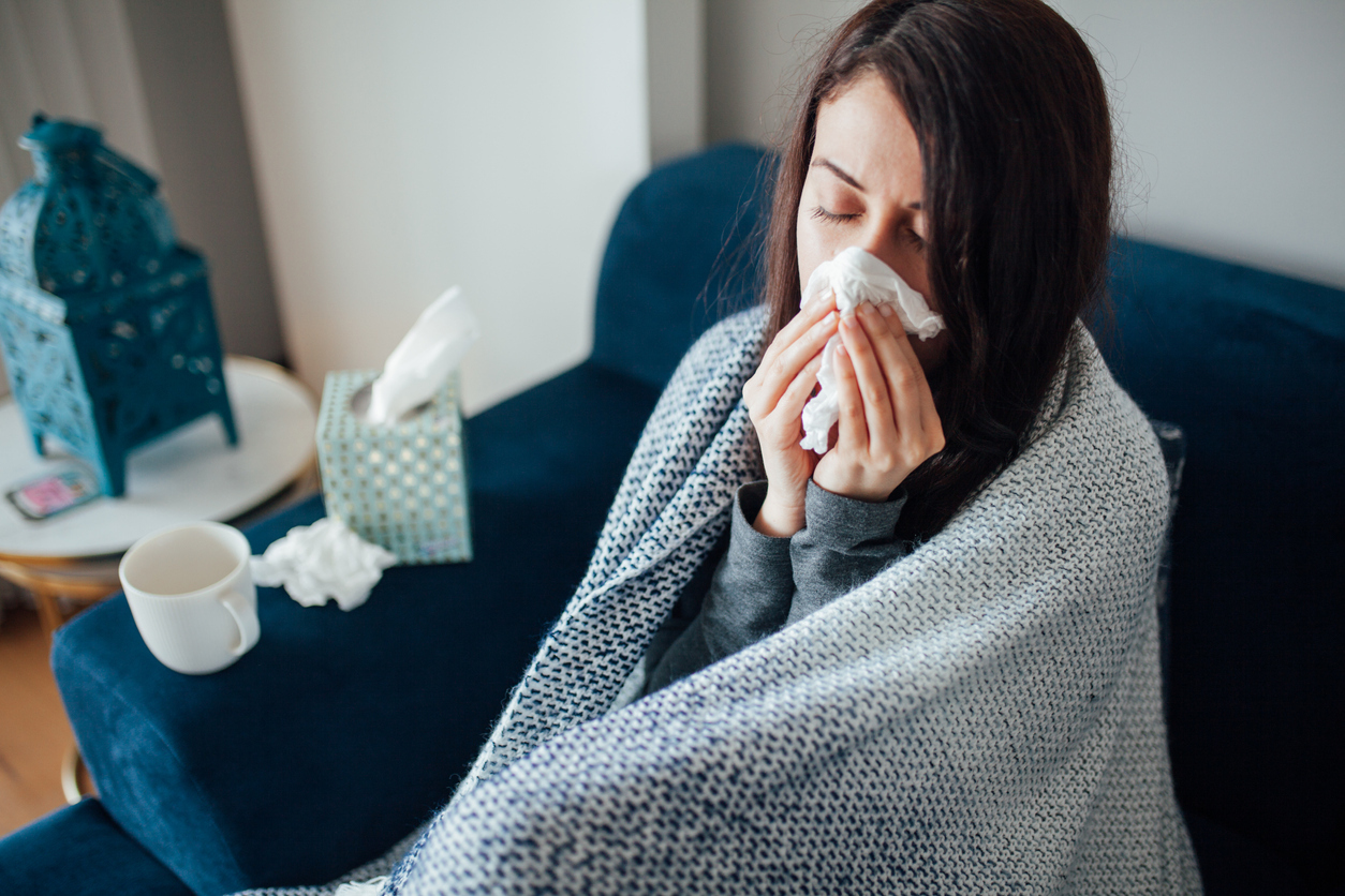 A young woman sitting on the couch wrapped in a blanket and blowing her nose, suffering from flu symptoms.