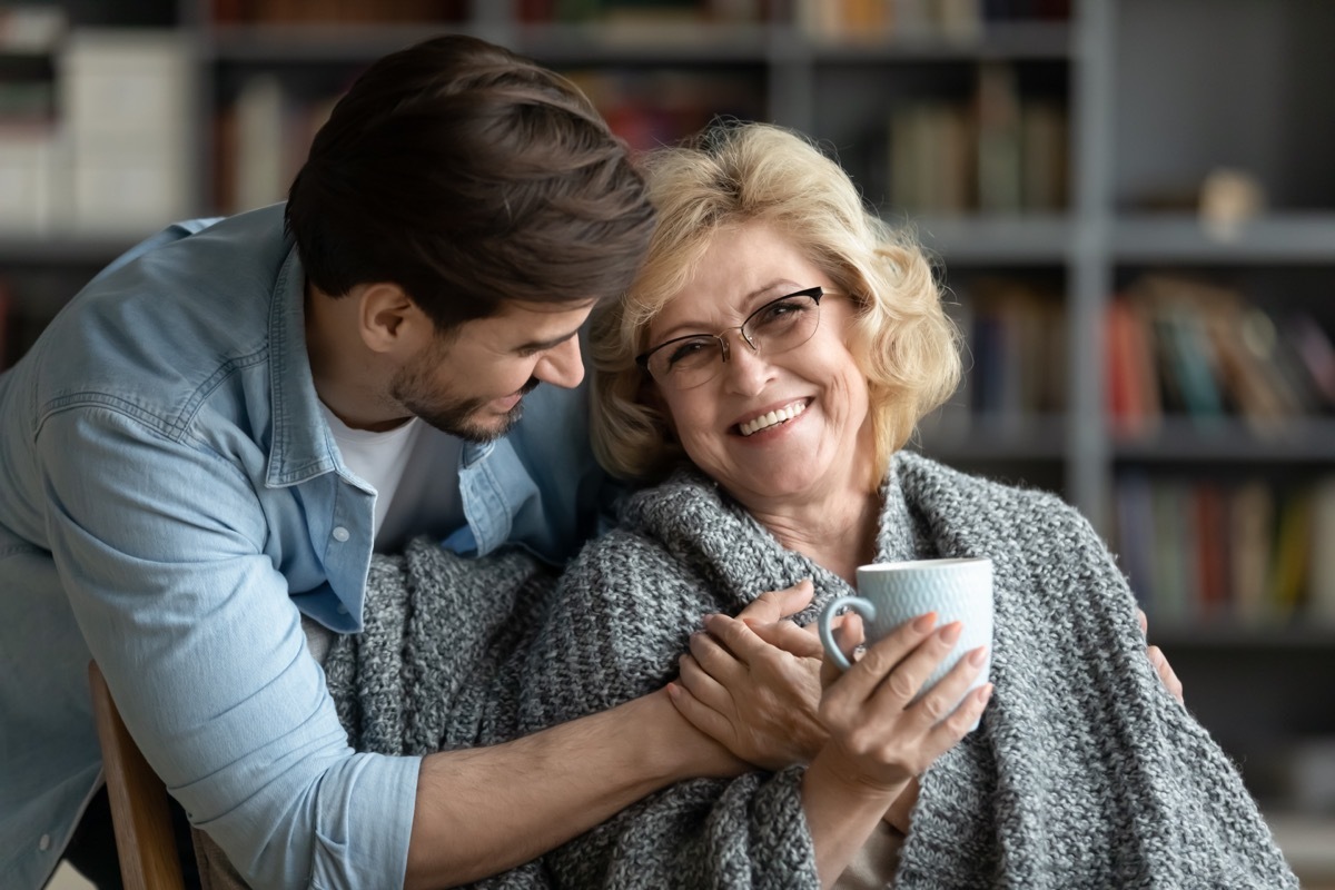 Happy middle-aged mother relax in chair drink tea enjoy family weekend reunion with grown-up son, smiling senior 70s mom rest at home spend time with caring adult man child, bonding concept