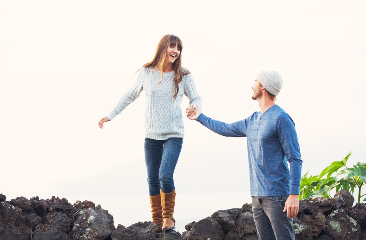 man holding woman's hand as she walks along stone wall