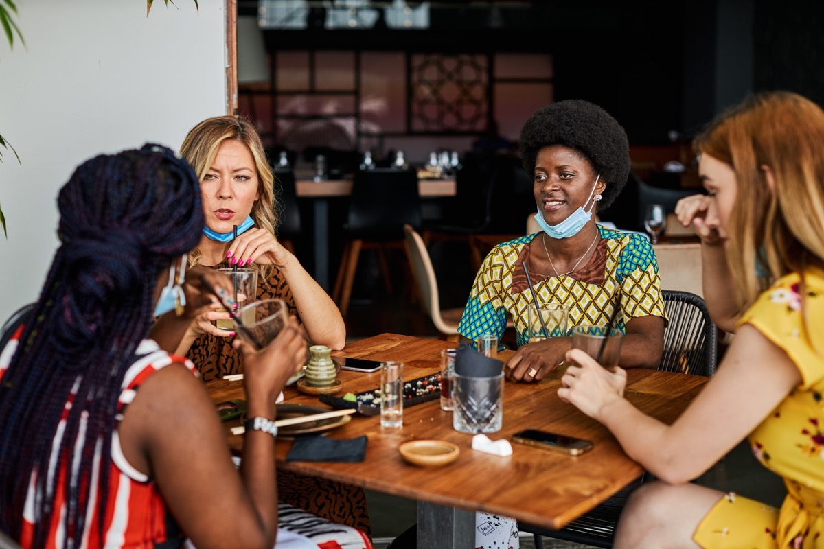 Four young women sitting in restaurant, wearing face mask on chin.