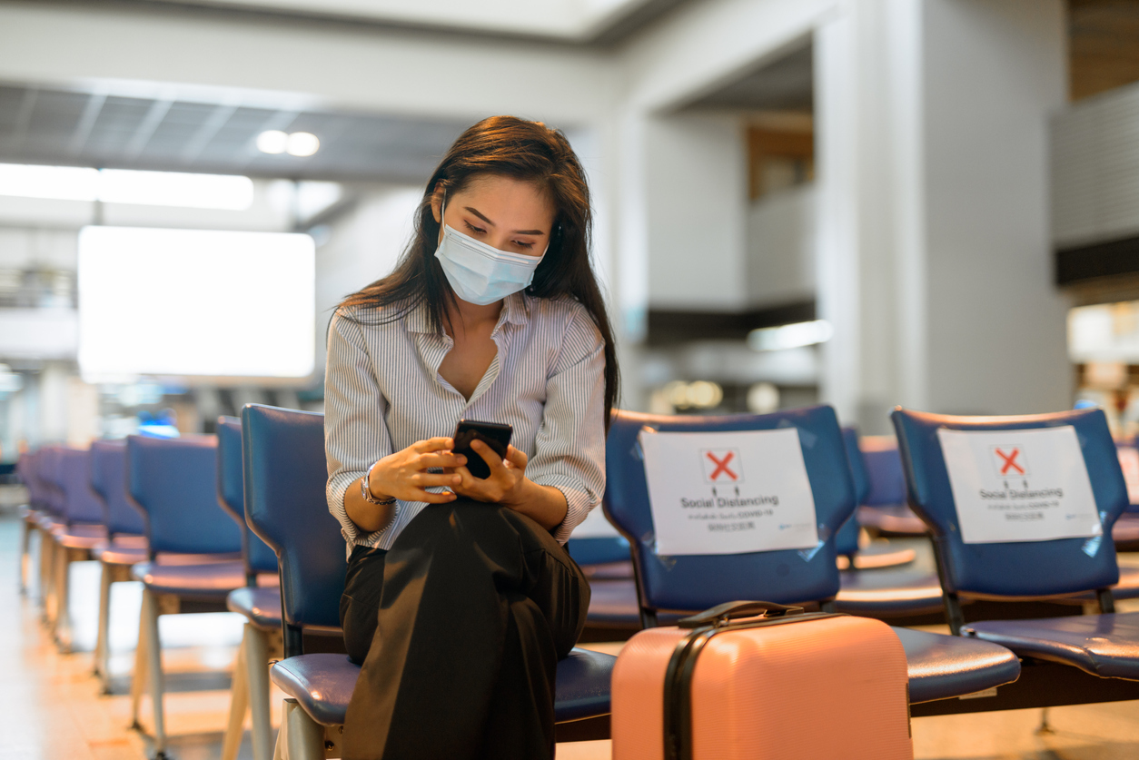 A young woman sitting in an airport while wearing a face mask, sitting next to socially distanced seats