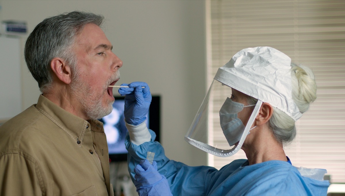 A mature Caucasian man in a clinical setting being swabbed by a healthcare worker in protective garb to determine if he has contracted the coronavirus or COVID-19.