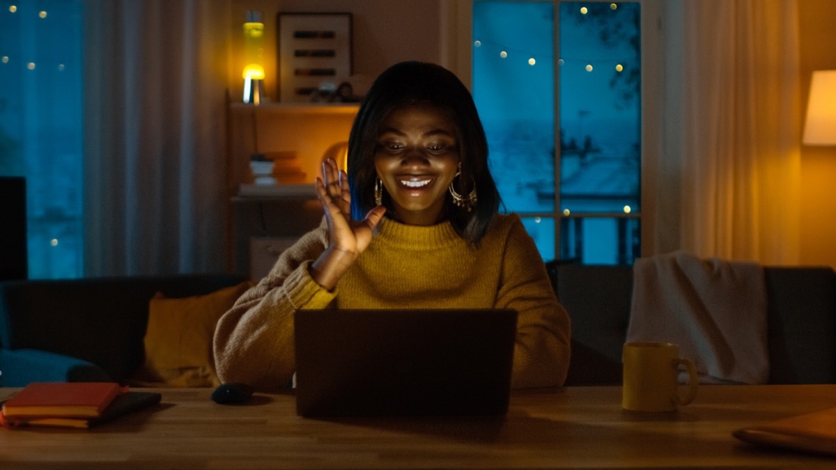 young black woman waving at her computer while videochatting at night