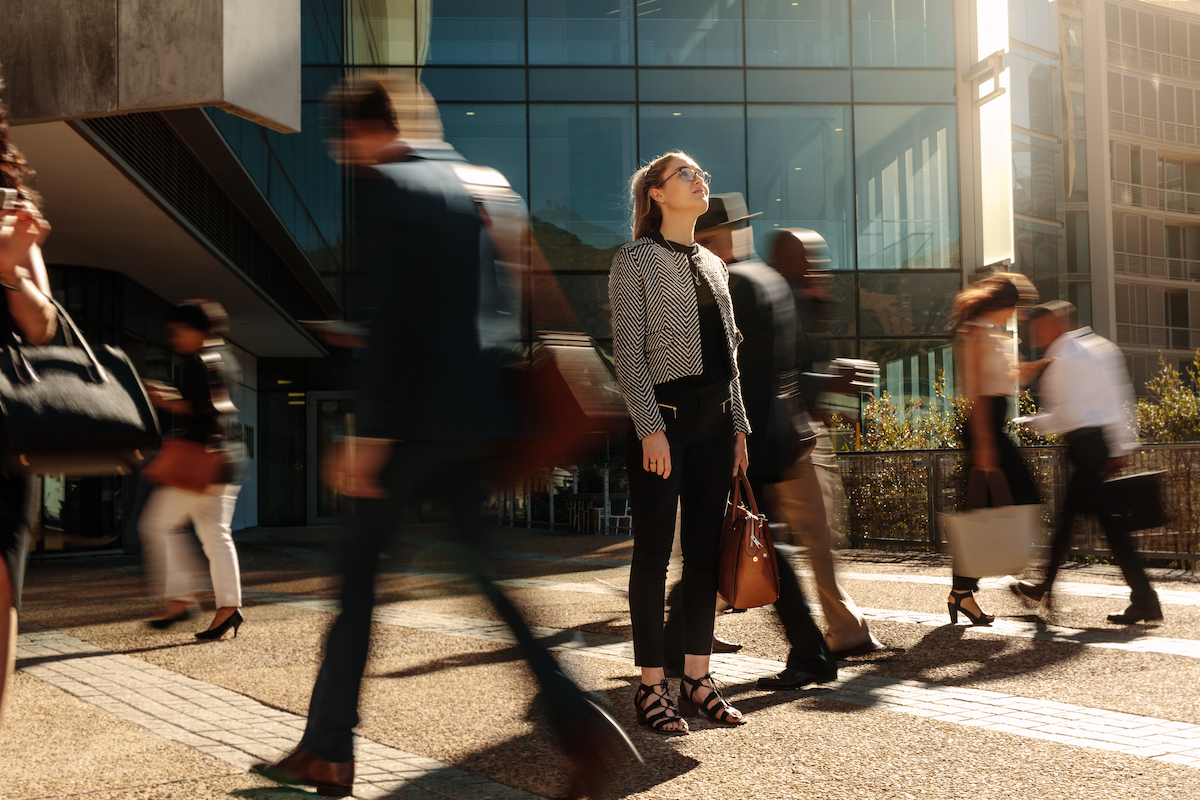 Woman standing amidst a busy office going crowd hooked to their mobile phones. Businesswoman holding her hand bag standing still on a busy street with people walking past her using mobile phones.