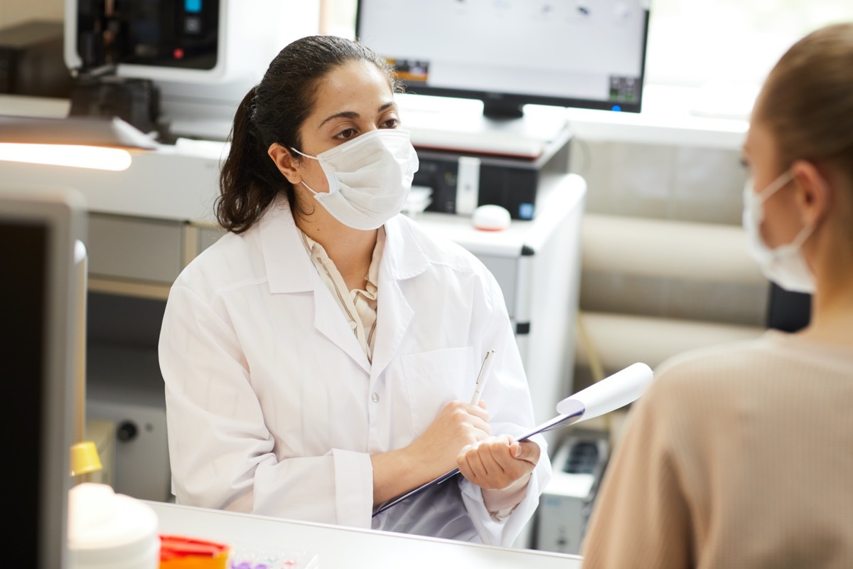 Female doctor in mask making notes in medical card while talking to patient at hospital