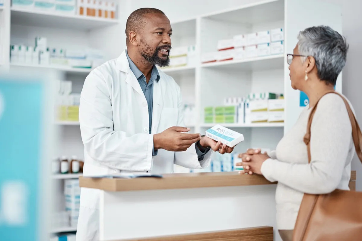 Healthcare, pharmacist and woman at counter with medicine or prescription drugs in hands at drug store. Health, wellness and medical insurance, man and customer at pharmacy for advice and pills.