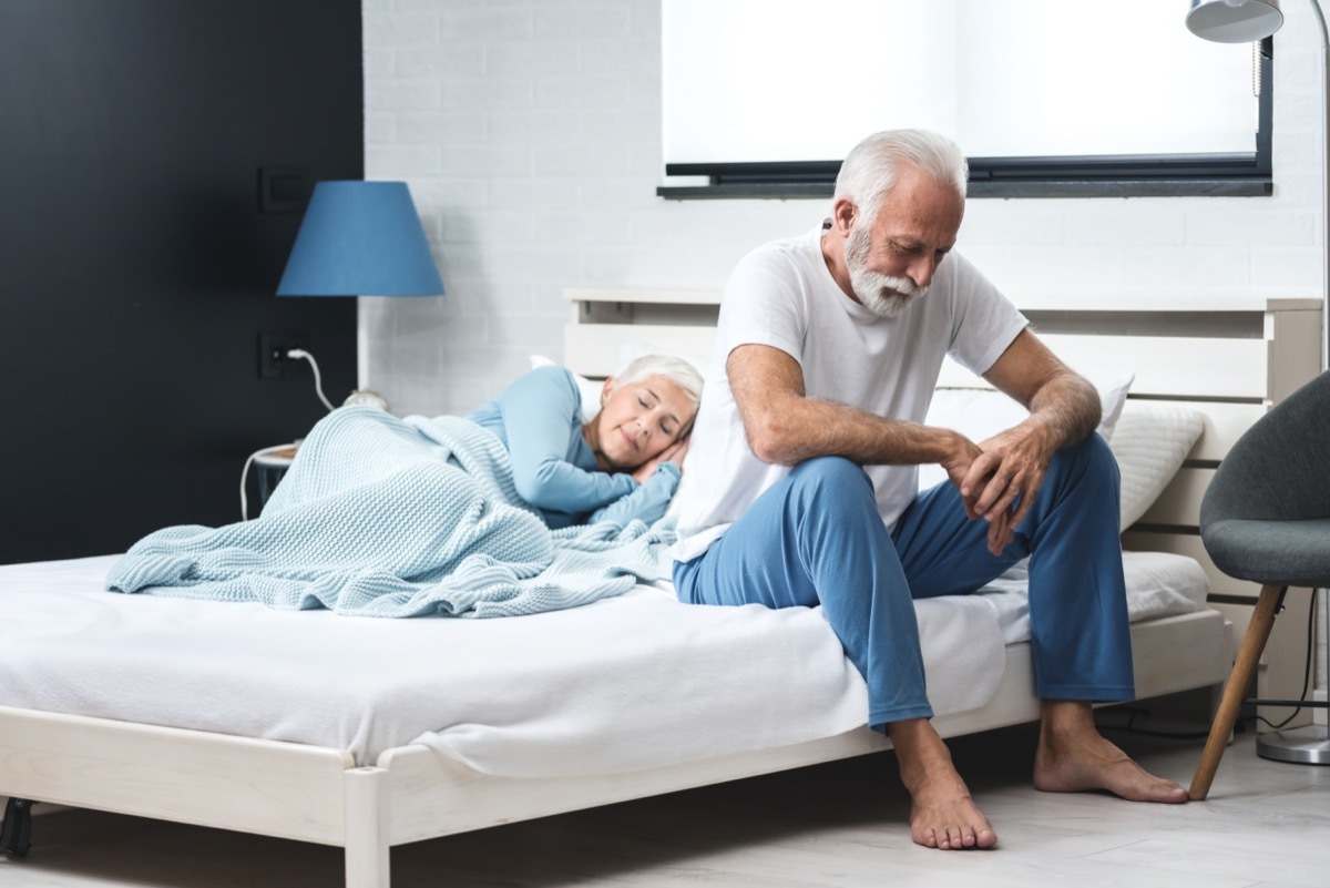 Worried senior depressed man sitting in bed and suffering from insomnia while his wife sleep