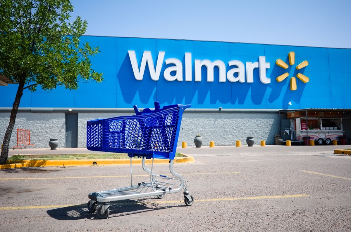 Shopping cart on a parking lot in front of main entrance to Walmart supermarket outdoor on the street with no people. Big Walmart logo on blue background behind.