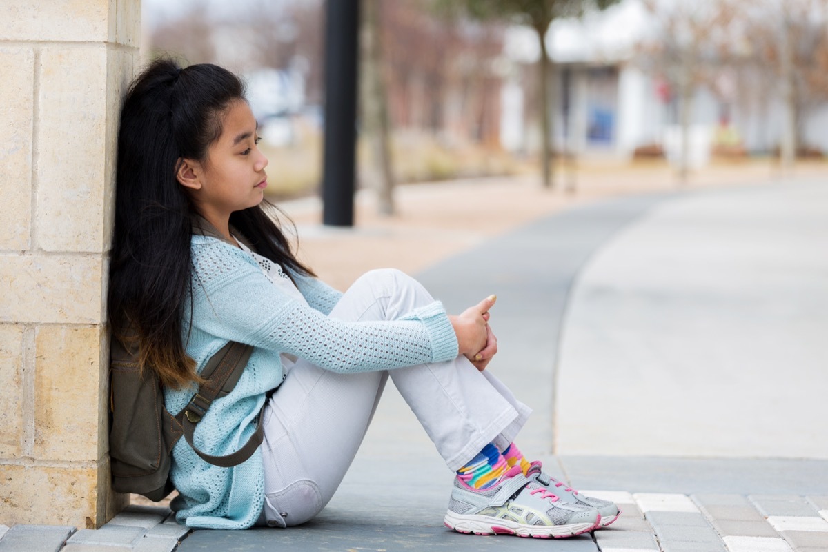 sad girl sitting outside on the sidewalk of her elementary school by herself