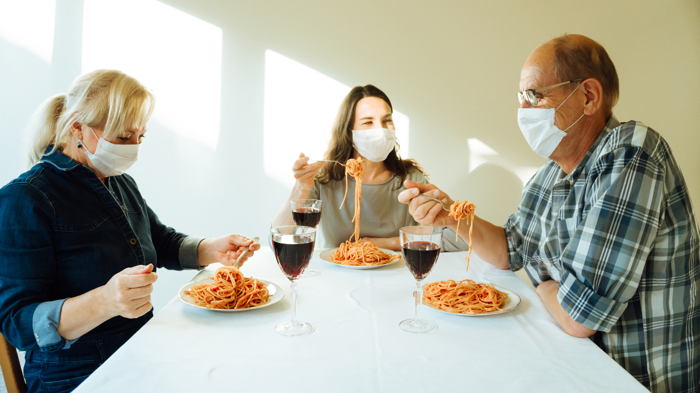 Three family members sit together while eating spaghetti and wearing face masks.