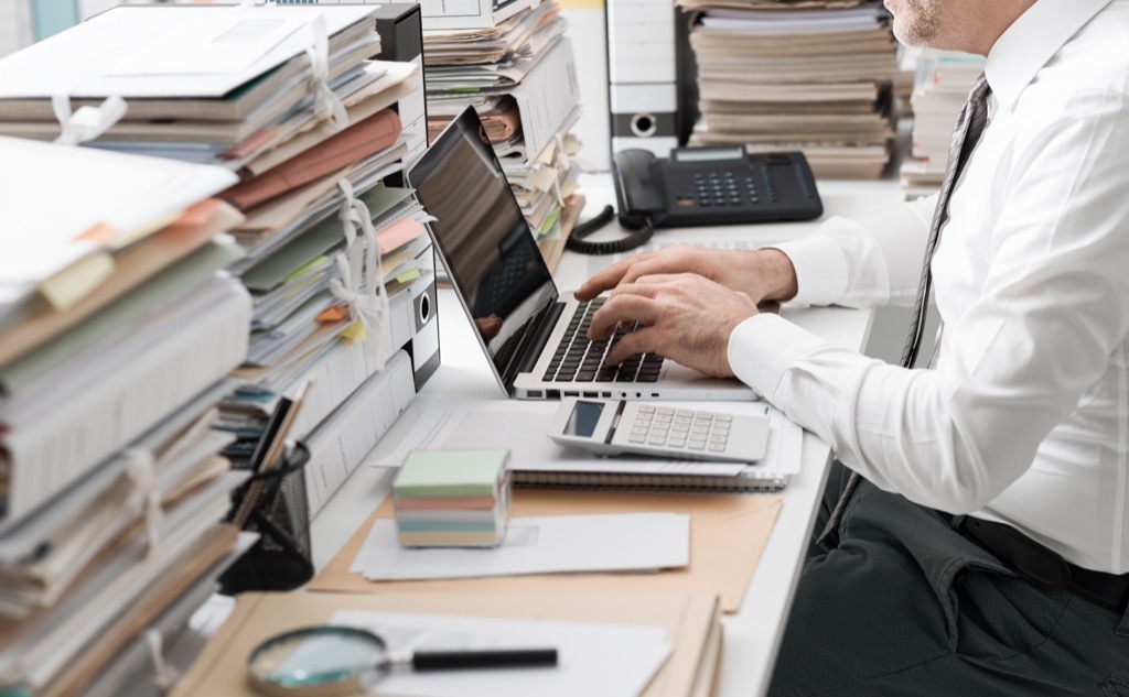 Frustrated businessman at the computer with stacks of paper.