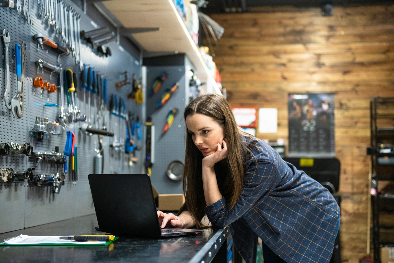 A woman using a laptop on a workbench with tools hanging