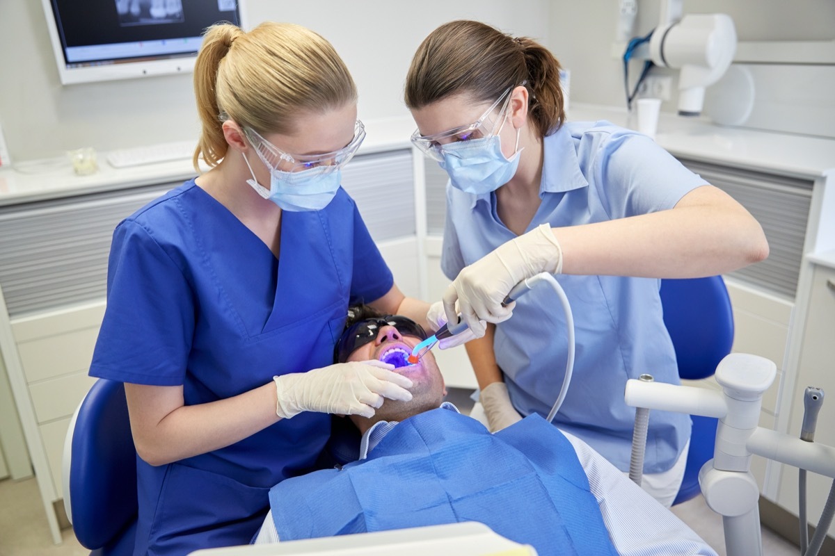 female dentist and assistant with dental curing light and mirror treating male patient teeth at dental clinic office