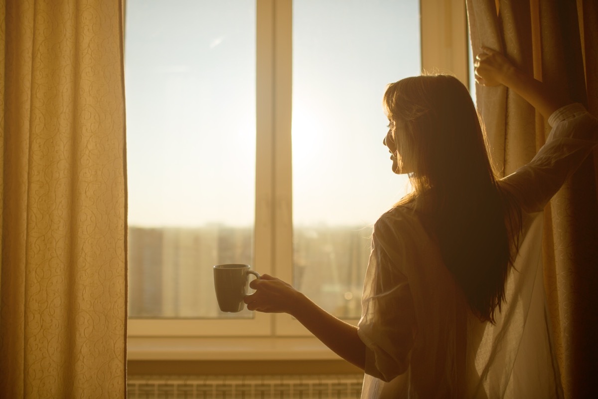 Woman opening curtains in morning with coffee