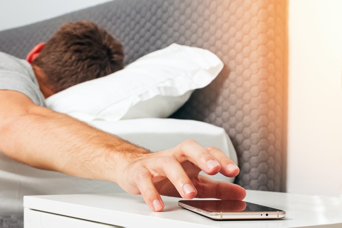 Man's hand close-up trying to reach out ringing cell phone or alarm to have some snooze early in the morning.