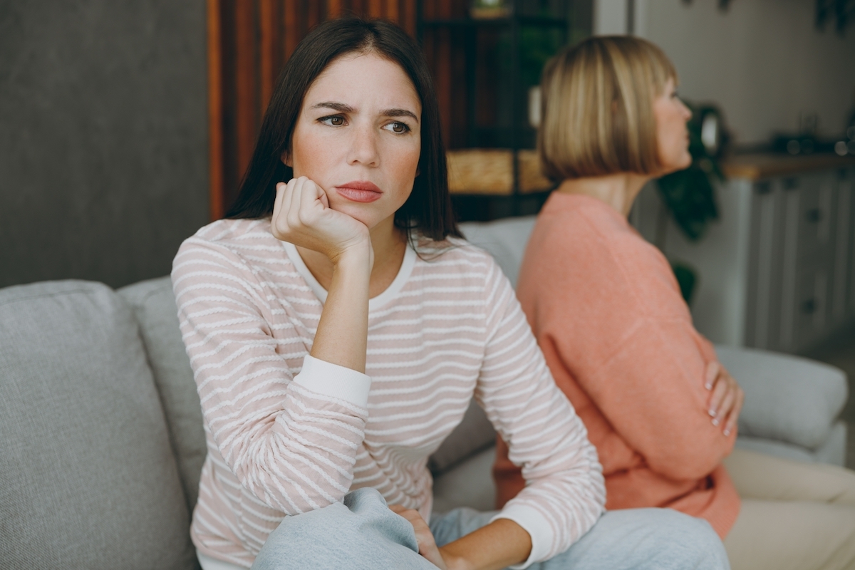 Two sad pensive depressed unhappy upset adult women mature mom young kid wearing casual clothes sit on gray sofa couch stay at home flat rest relax spend free spare time in living room. Family concept