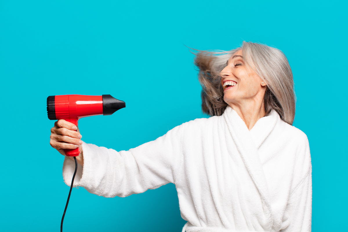 An older woman with gray hair in a white robe using a blow dryer against a teal background.