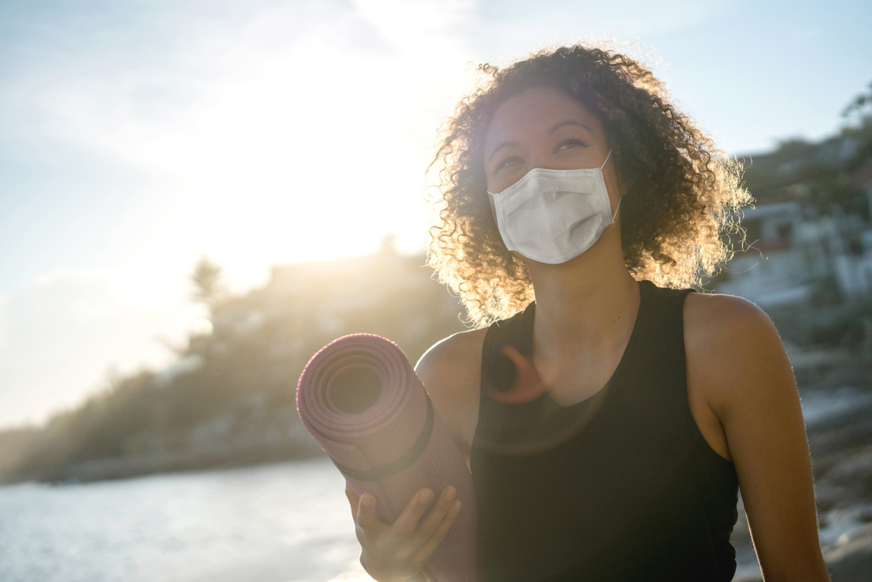 A young woman wearing a face mask outdoors while holding a yoga mat.