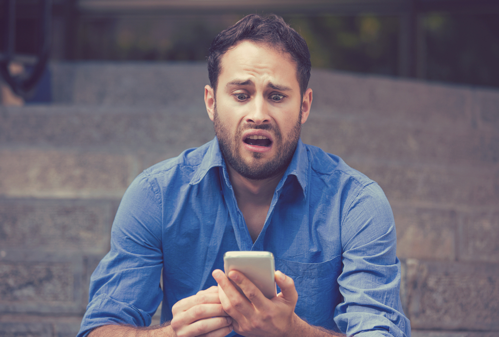 A young man looking at his smartphone with a horrified look on his face.