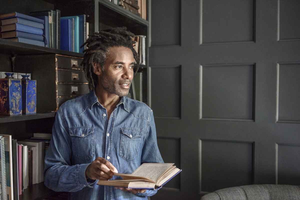 middle aged black man holding open a book next to his bookshelf