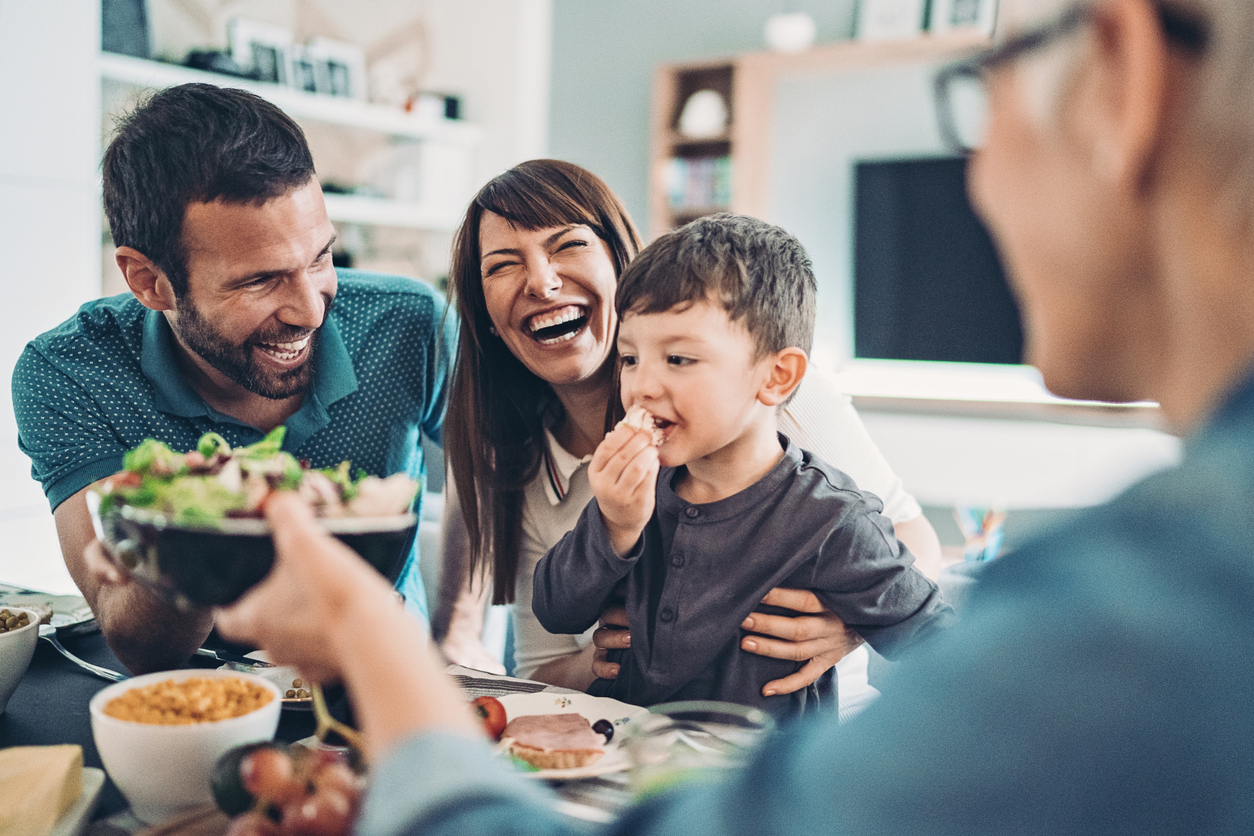 A mother, father, and son sit laughing at the dinner table while a grandmother passes them a bowl of salad