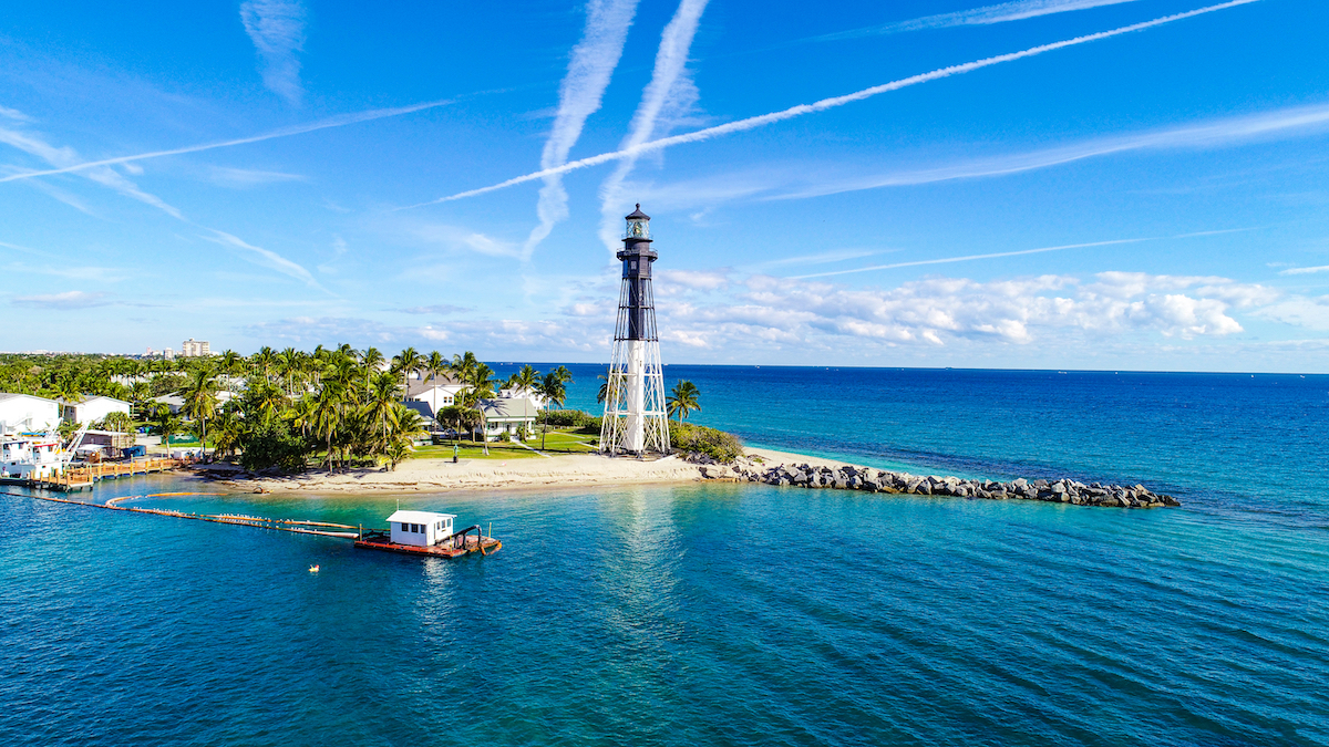 The Hillsboro Inlet Lighthouse in Fort Lauderdale, Florida. The black and white structure is set on a sandy shore with the teal-colored ocean.