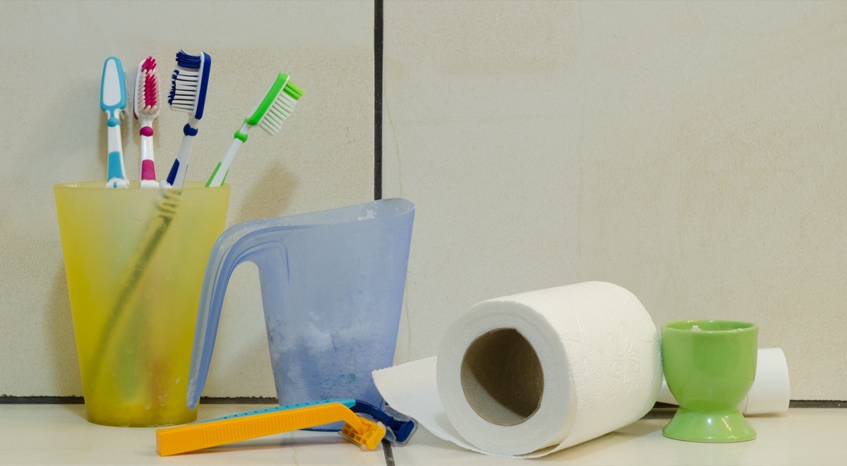 Messy toilet shelf with a blue cup, yellow cup with toothbrushes, toilet paper, green cup and two razors