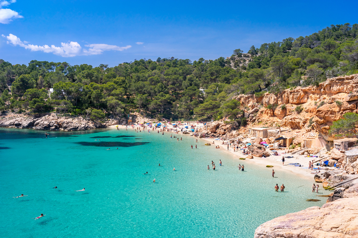 Wide-angle view of Cala Saladeta, a few steps from Cala Salada, on the western coast of Ibiza, renowned for its extremely transparent waters, pearly sands and a scenic group of fishermen's huts (locally known as casitas varadero). Bathers engaged in different activities, the bright light of a summer noon, picturesque clouds, colourful cliffs, lush vegetation. 